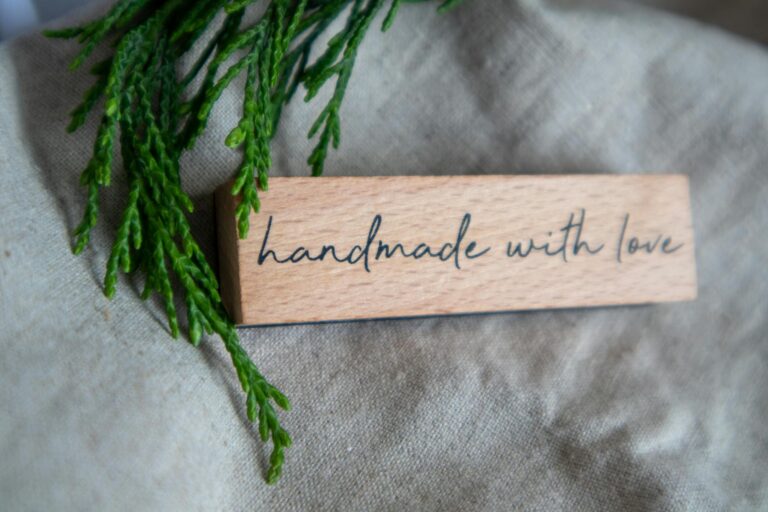 A rustic wooden sign with 'Handmade with Love' text on linen, accented by green foliage.